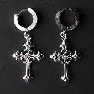 May include: A pair of silver-colored hoop earrings with ornate cross pendants. The crosses have a detailed, gothic design with decorative flourishes and a small gem in the center. The earrings are displayed against a dark background.