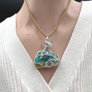 May include: A pendant necklace featuring a turquoise stone with brown inclusions, wrapped in silver-toned wire. The wire design incorporates two dolphin figures and a small shell-like accent. The pendant hangs from a tan suede cord necklace.