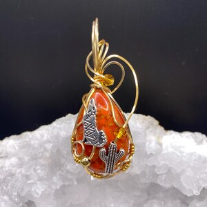 May include: A teardrop-shaped pendant with a red and orange stone, wrapped in gold wire. It features silver charms of a howling wolf and a cactus. The pendant is displayed on a white, textured surface.