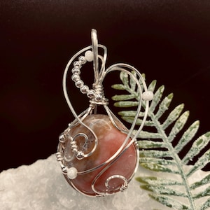 May include: A handcrafted pendant with a polished, reddish-brown gemstone encased in intricate silver wire. The wirework creates a decorative frame around the stone, with small silver beads and accents. The pendant is displayed on a white surface.