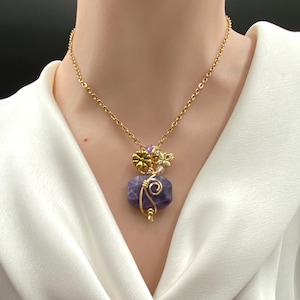 May include: A gold-colored chain necklace with a pendant featuring a purple gemstone. The pendant is adorned with gold-colored wire, a small gold flower, and a butterfly. The necklace is worn against a white background.