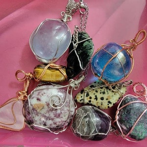 Op de afbeelding: Een verzameling van acht draadgewikkelde edelsteen hangers. De hangers bevatten een verscheidenheid aan stenen, waaronder amethist, tijgeroog, rozenkwarts, dalmatijnse jaspis en een blauwe steen. De hangers zijn allemaal gewikkeld in koperdraad en hebben een rustieke, handgemaakte uitstraling.