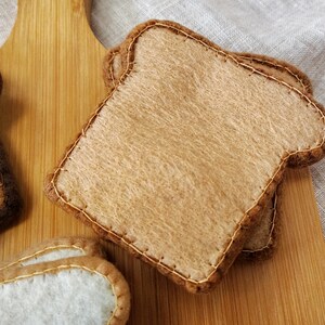 May include: Two slices of felt toast with brown edges and tan centers, stitched with gold thread. The toast is on a wooden cutting board. Other felt food items are visible.