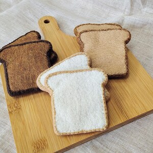 May include: A wooden cutting board with four felt bread slices. Two slices are light brown, two are white, and all are stitched with contrasting thread. The bread slices are arranged on the board.