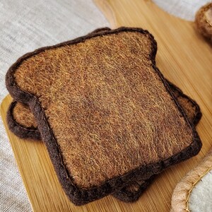 May include: Two slices of felt toast with brown crusts and textured, tan interiors, resting on a wooden cutting board. The felt bread has a handmade appearance, suggesting a craft or toy item.