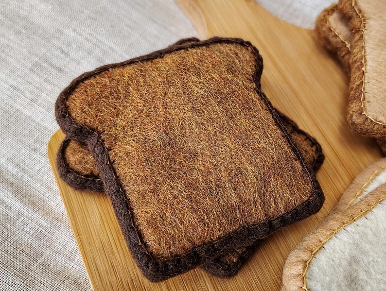 May include: Two slices of felt toast with brown crusts and textured, tan interiors, resting on a wooden cutting board. The felt bread has a handmade appearance, suggesting a craft or toy item.