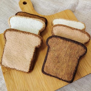 May include: A wooden cutting board holds several felt bread slices in various shades of brown and white. The bread slices are arranged to show different levels of toasting, with some appearing lightly toasted and others deeply browned.