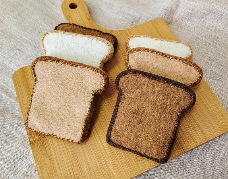 May include: A wooden cutting board holds several felt bread slices in various shades of brown and white. The bread slices are arranged to show different levels of toasting, with some appearing lightly toasted and others deeply browned.