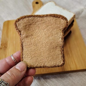 May include: A close-up of a felt bread slice with brown crust and tan interior, stitched with brown thread. The bread is on a wooden cutting board with other slices. The image suggests a food-themed craft or toy.