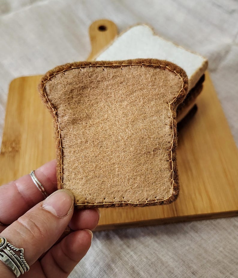 May include: A close-up of a felt bread slice with brown crust and tan interior, stitched with brown thread. The bread is on a wooden cutting board with other slices. The image suggests a food-themed craft or toy.