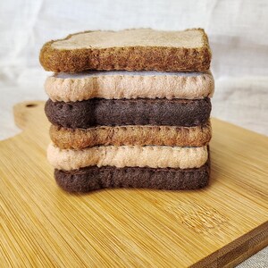 May include: A stack of felt sandwiches with layers of brown, tan, and beige. The sandwich is on a wooden cutting board. The bread is a darker brown, and the fillings are lighter shades of brown and tan.