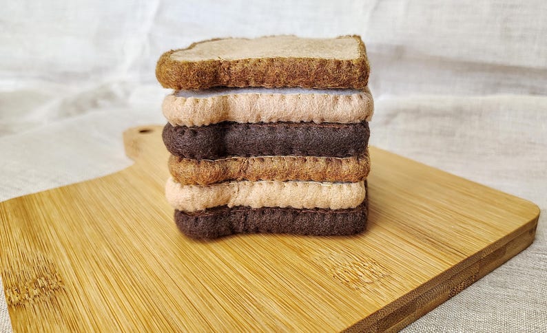 May include: A stack of felt sandwiches with layers of brown, tan, and beige. The sandwich is on a wooden cutting board. The bread is a darker brown, and the fillings are lighter shades of brown and tan.