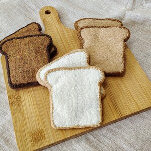 May include: A wooden cutting board with several felt bread slices. The bread slices are in various shades of brown and white, with stitched edges. The cutting board is made of light-colored wood.