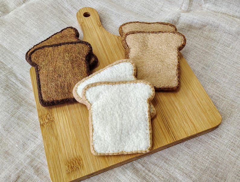 May include: A wooden cutting board with several felt bread slices. The bread slices are in various shades of brown and white, with stitched edges. The cutting board is made of light-colored wood.