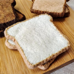 May include: Close-up of felt bread slices, including white bread and toasted brown bread, arranged on a wooden cutting board. The bread slices have gold stitching around the edges. The background is a light-colored fabric.