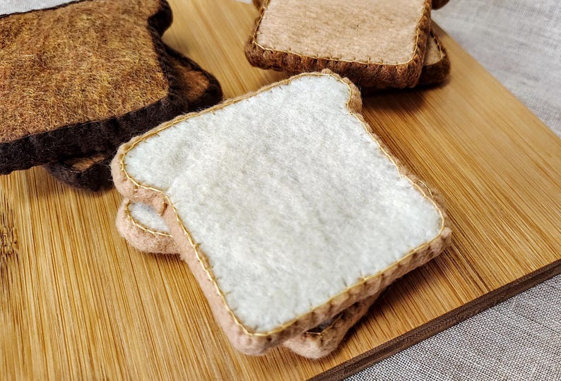 May include: Close-up of felt bread slices, including white bread and toasted brown bread, arranged on a wooden cutting board. The bread slices have gold stitching around the edges. The background is a light-colored fabric.
