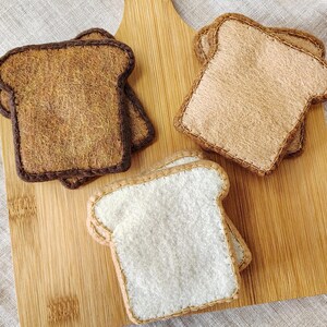 May include: Felt bread slices in various shades of brown and white, arranged on a wooden cutting board. The bread slices are stitched around the edges, with two slices of toasted bread and one slice of white bread.