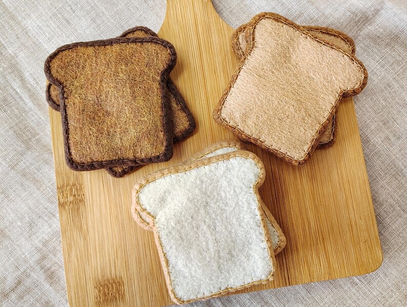 May include: Felt bread slices in various shades of brown and white, arranged on a wooden cutting board. The bread slices are stitched around the edges, with two slices of toasted bread and one slice of white bread.