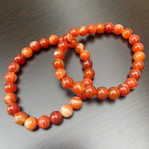 Puede incluir: Dos pulseras hechas con cuentas de cornalina naranja y blanca. Las pulseras est&aacute;n apiladas una encima de la otra.