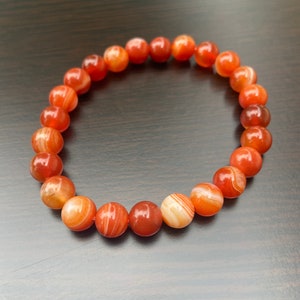 Puede incluir: Un brazalete de cornalina roja con rayas blancas. El brazalete est&aacute; hecho de cuentas redondas y tiene una longitud aproximada de 18 cm.