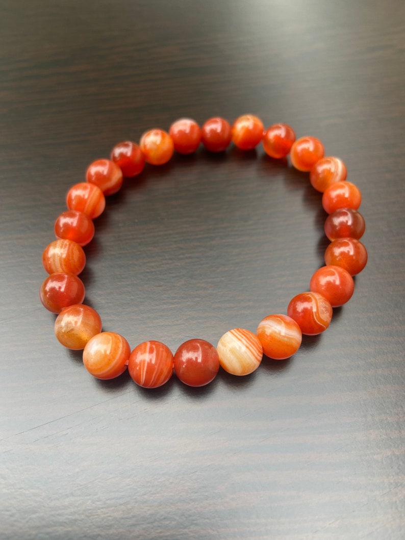 Puede incluir: Un brazalete de cornalina roja con rayas blancas. El brazalete est&aacute; hecho de cuentas redondas y tiene una longitud aproximada de 18 cm.