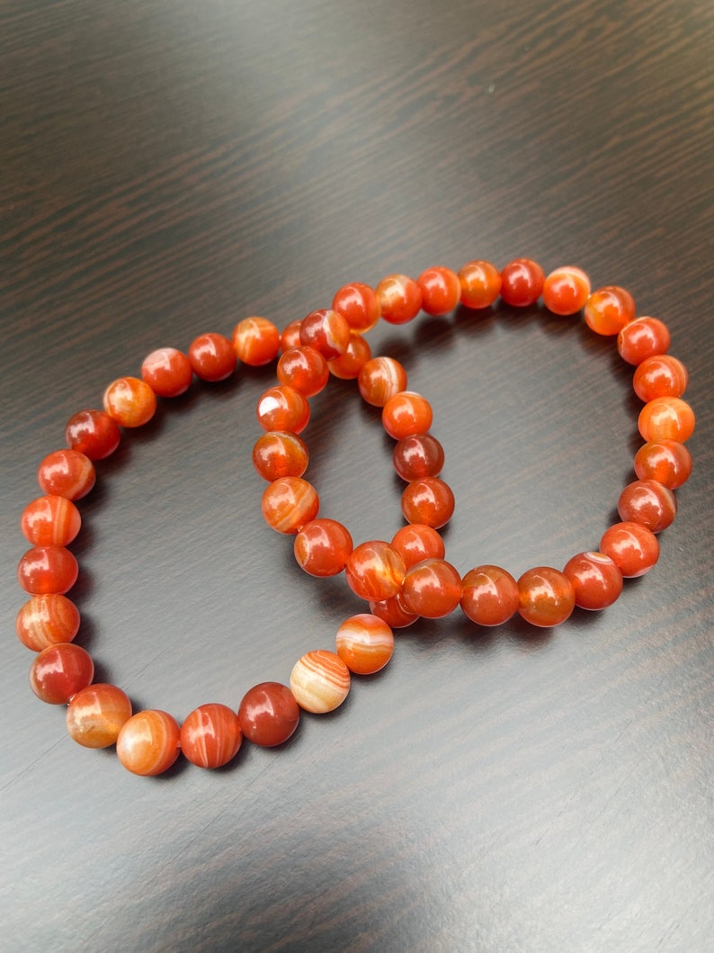 Puede incluir: Dos pulseras hechas con cuentas de cornalina naranja y blanca. Las pulseras est&aacute;n apiladas una encima de la otra.