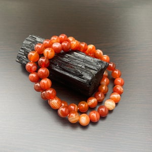 Puede incluir: Un brazalete de doble cadena hecho con cuentas de cornalina. Las cuentas son de color naranja intenso con rayas blancas. El brazalete est&aacute; apoyado sobre una piedra de obsidiana negra.
