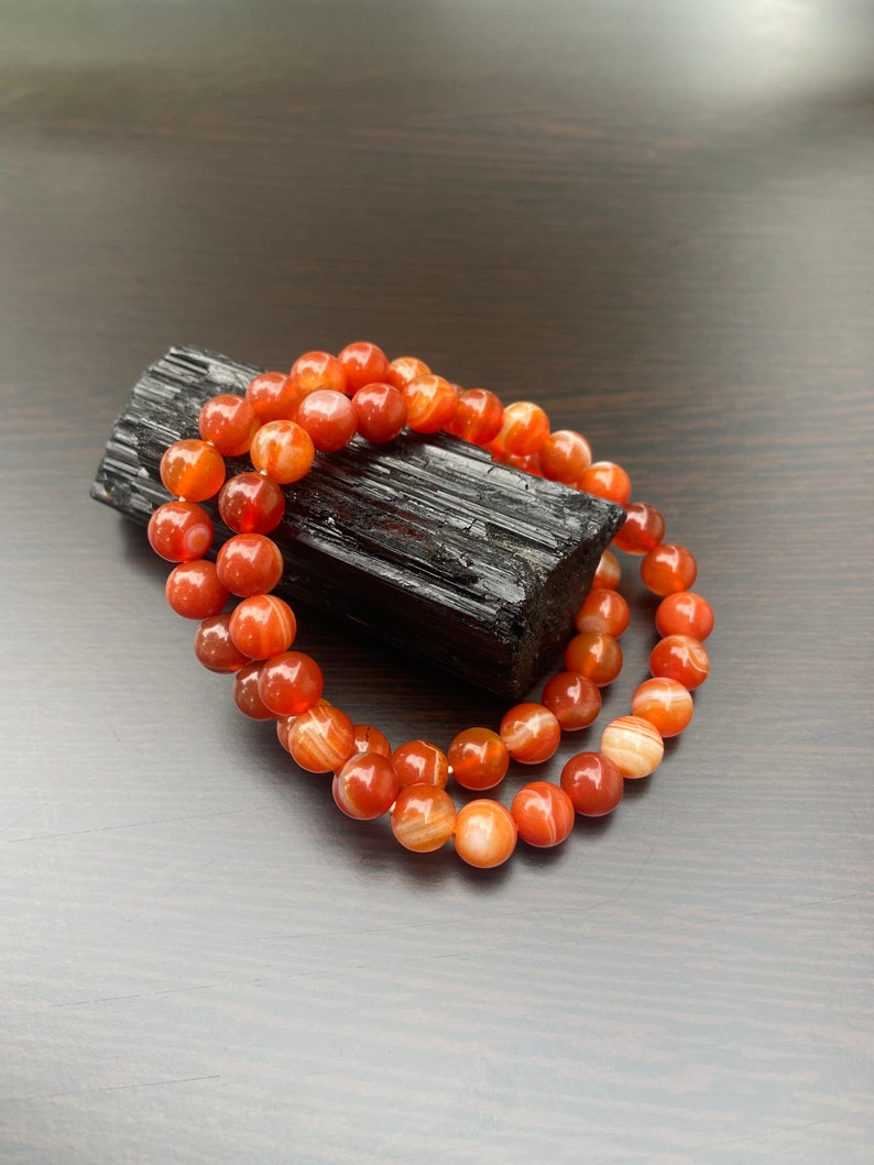Puede incluir: Un brazalete de doble cadena hecho con cuentas de cornalina. Las cuentas son de color naranja intenso con rayas blancas. El brazalete est&aacute; apoyado sobre una piedra de obsidiana negra.