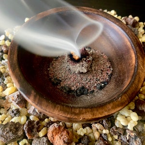 Puede incluir: Un cuenco de madera con un acabado marr&oacute;n oscuro, lleno de una mezcla gris y roja, con humo que sale del centro. El cuenco est&aacute; rodeado de peque&ntilde;as piedras de color marr&oacute;n claro y amarillo.