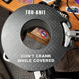 May include: A circular, textured, dark gray device with the text "TRU-KNIT" and "DON'T CRANK WHILE COVERED" in white. The device has a central opening and a silver-colored handle. The item appears to be a tool or part of a larger machine.