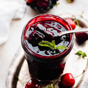 Puede incluir: Un tarro de mermelada de cerezas oscuras con una ramita de menta, sobre una bandeja plateada. Las cerezas frescas están esparcidas alrededor del tarro y en un pequeño cuenco negro. Una cuchara plateada descansa en la mermelada, de color rojo oscuro.