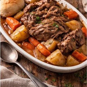 Puede incluir: Un plato abundante de carne de res estofada con zanahorias y patatas en una fuente ovalada blanca. Un panecillo está al lado. Una cuchara de plata descansa sobre una servilleta de lino, con una superficie de madera en el fondo.