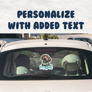 May include: A white car with a rear window sticker featuring a brown dog with a blue collar and a flower crown. The sticker also includes the name "Mabel" in teal text.