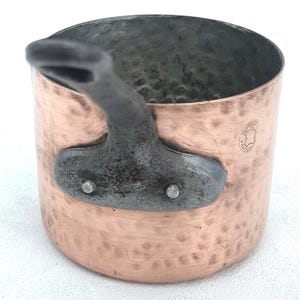 May include: A hammered copper saucepan with a dark, textured metal handle attached by two rivets. The exterior surface features a distinct hammered finish and a small circular stamp that reads Fabrication Francaise, indicating its origin.