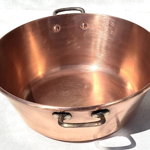 Puede incluir: Un cuenco redondo de cobre pulido con una abertura ancha y una superficie interior lisa y reflectante. El recipiente cuenta con dos asas robustas de metal oscuro fijadas a los lados con remaches visibles para facilitar su agarre.