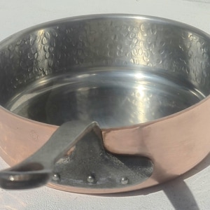 Puede incluir: Una sartén redonda y poco profunda con un exterior de cobre pulido y un interior de acero inoxidable martillado. La sartén cuenta con un mango de metal oscuro y resistente, fijado al lateral con tres remaches visibles para un agarre firme.