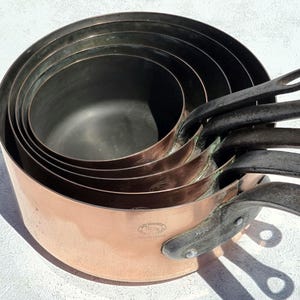 Könnte beinhalten: Ein ineinander gestapeltes Set aus fünf runden Kupferkasserollen mit dunklen Metallgriffen. Jede Pfanne hat eine polierte Kupferaußenseite und eine dunkle Innenseite. Die größte Pfanne zeigt einen runden Stempel Mauviel Made in France.