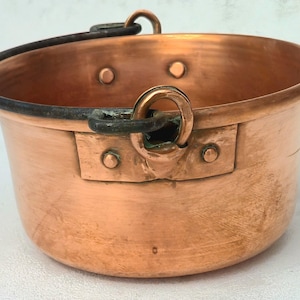 Puede incluir: Una olla redonda de cobre con un acabado metálico pulido, que cuenta con un asa giratoria de hierro oscuro y desgastado, sujeta a los lados con soportes metálicos remachados. El recipiente tiene una parte superior abierta y un cuerpo curvo.