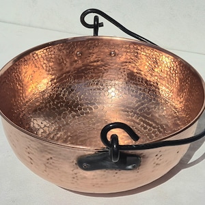 May include: A round, hammered copper bowl featuring a polished metallic finish and a textured surface. The bowl includes a black metal swing handle attached to two black metal brackets on opposite sides of the rim, secured with visible rivets.