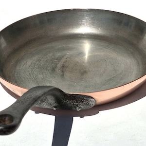 Op de afbeelding: Een ronde koekenpan met een gepolijste koperen buitenkant en een metallic zilverkleurig binnenoppervlak. De pan is voorzien van een donker, getextureerd ijzeren handvat dat met twee zichtbare klinknagels aan de zijkant is bevestigd.