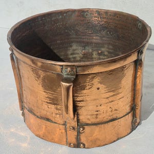 Op de afbeelding: Een grote, cilindervormige koperen bak met een gehamerde textuur, versterkt met verticale metalen banden en klinknagels. De buitenkant heeft een warme, metalen patina, terwijl de binnenkant ingewikkelde, herhalende cirkelvormige patronen toont.