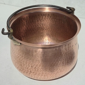 May include: A round, hammered copper pot featuring a wide, open top and a dark metal swing handle attached to the sides with brass-coloured hardware. The interior and exterior surfaces display a consistent, textured, dimpled pattern throughout.