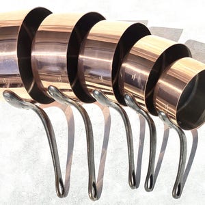 May include: A set of five graduated copper saucepans with polished metallic exteriors and dark, curved iron handles. The pans are arranged in a row from largest to smallest, each featuring a cylindrical body and a sturdy, riveted handle.
