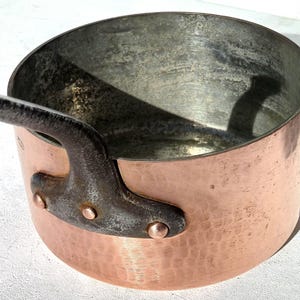 Puede incluir: Un cazo redondo de cobre con acabado exterior martillado y revestimiento interior metálico. El cazo cuenta con un mango de hierro oscuro y texturizado, fijado al lateral con tres remaches de cobre, mostrando signos de uso y pátina.