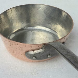 Może przedstawiać: Okrągły miedziany rondel z młotkowanym wykończeniem zewnętrznym i gładką, metaliczną powłoką wewnętrzną. Rondel posiada solidny, nitowany metalowy uchwyt przymocowany z boku, kontrastujący ciepły odcień miedzi ze srebrzystym kolorem uchwytu.