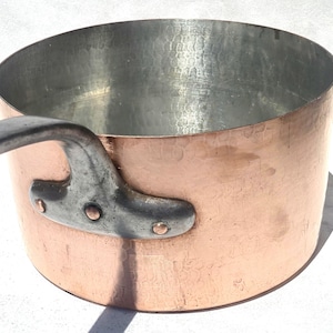 Op de afbeelding: Een koperen steelpan met een zilveren handvat bevestigd met klinknagels. De binnenkant van de pan is zilverkleurig en de buitenkant heeft een warme koperkleur. De pan is rond en heeft een gehamerde textuur.