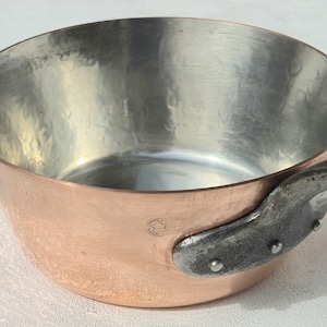 May include: A round copper saucepan with a polished silver-toned interior and a dark, textured metal handle attached with three visible rivets. The exterior copper surface features a small circular maker's mark near the handle attachment point.
