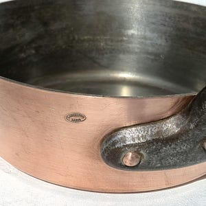 Puede incluir: Una cacerola de cobre con interior oscuro y asa de hierro remachada. La cacerola tiene forma redondeada y exterior de cobre brillante. Un pequeño emblema ovalado con texto es visible en el lateral.