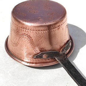 May include: A copper ladle with a hammered finish and a dark metal handle. The ladle has a rounded bowl shape with decorative patterns near the rim. The handle is attached with rivets.