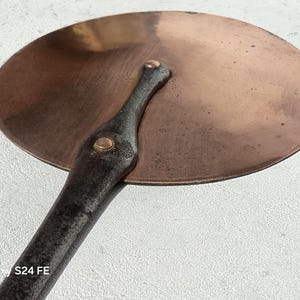 Peut inclure: Un couvercle de cuisson rond de couleur cuivrée avec une longue poignée sombre. Le couvercle a une forme légèrement bombée et est fixé à la poignée par des rivets. La poignée est sombre et texturée, suggérant qu'elle est en fer. L'aspect général est rustique et vintage.